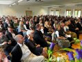 Menschen sitzen auf Stühlen in Reihen in einem vollen Saal.