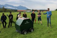 Menschen stehen um einen grünen Metallkasten auf Rädern auf einer Wiese.