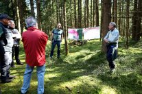 Zwei Förster stehen neben zwischen zwei Bäume gespannten Plakaten und erklären etwas