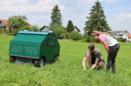 ein grüner, kastenförmiger Roboter, daneben kniet ein Mann und zeigt einer Frau etwas im Gras