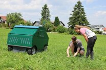 ein grüner, kastenförmiger Roboter, daneben kniet ein Mann und zeigt einer Frau etwas im Gras